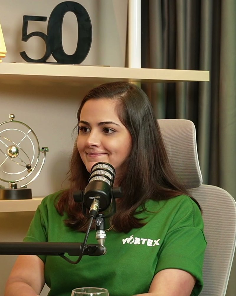 Carol Fonseca sorrindo, ao microfone, na mesa do estúdio.