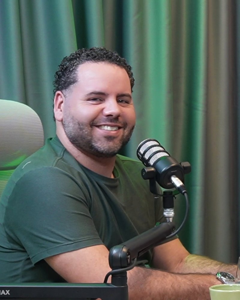 Léo Godoy sorrindo, ao microfone, na mesa do estúdio.